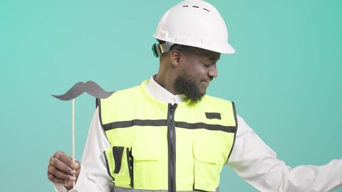 الإنشاءات المعمارية في الأراضي السعودية، المحافظة على اللياقة والقوة العضلية، اليوم العالمي للرجل 19 نوفمبر، العمل في القطاع الهندسي، بورتريه لمهندس أسمر عربي خليجي سعودي مبتسم يرتدي الخوذه و سترة الحماية و يمسك بيده عود خشبي مع قصاصة ورقية على شكل شارب و يستعرض عضلاته، خلفية زرقاء