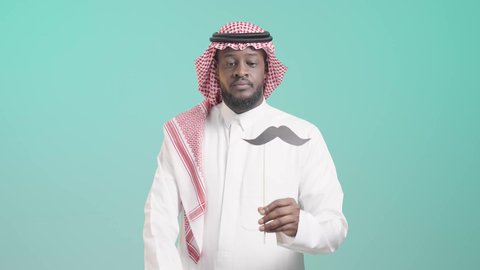 طلب التوقف عن فعل شيء ما، رفع اليد بايماءات التحذير والتنبيه، الإهتمام بصحة الذكور، اليوم العالمي للرجل 19 نوفمبر، الإحتفال بالمناسبات العالمية، بورتريه لرجل أسمر عربي خليجي سعودي يرتدي الثوب التقليدي والشماغ يمسك بيده عود خشبي مع قصاصة ورقية على شكل شارب، خلفية زرقاء