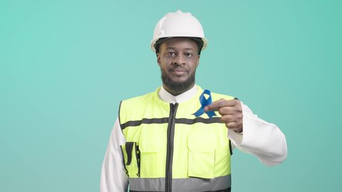 The concept of architectural construction and engineering, skill and mastery of work, working in the engineering sector, portrait of a smiling Saudi Arabian Gulf brown engineer wearing a helmet and a protective jacket and pointing with his index finger to a blue ribbon indicating high quality, Saudi professions and jobs, blue background