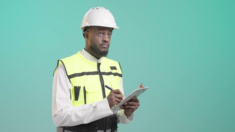 كتابة آخر المستجدات حول المشروع، التأكد من سير الأعمال بالشكل المطلوب والصحيح، بورتريه لمهندس أسمر عربي خليجي سعودي يرتدي الثوب التقليدي و السترة العاكسة و خوذة الحماية يمسك الملف بيده ويكتب فيه بإيماءات التركيز، خلفية زرقاء