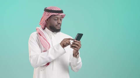 Using mobile phones and modern technology to facilitate daily tasks, browsing the internet and working remotely, portrait of a dark-skinned Saudi Arabian Gulf man wearing traditional thobe and shemagh and using a mobile phone, tapping on the mobile screen, blue background