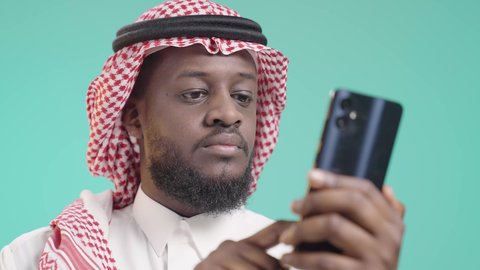 تبادل الأحاديث مع الأهل والأصدقاء عبر مواقع التواصل الاجتماعي، استخدام الجوال والتكنولوجيا الحديثة لتسهيل القيام بالمهام اليومية, تصفح الإنترنت والعمل عن بعد, بورتريه لرجل أسمر عربي خليجي سعودي يرتدي الثوب التقليدي و الشماغ و يستخدم الهاتف المحمول, النقر على شاشة الموبايل, خلفية زرقاء