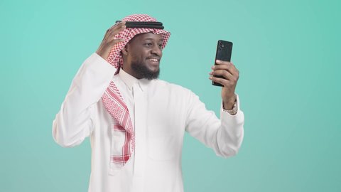 إجراء اتصال فيديو عبر الهاتف, التواصل مع الأهل والأصدقاء، دمج التكنولوجيا الحديثة في الحياة اليومية، بورتريه لرجل أسمر عربي خليجي سعودي مبتسم يرتدي الثوب التقليدي و الشماغ يتكلم عبر الهاتف المحمول, خلفية زرقاء