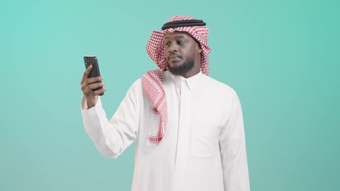Keeping up with technical development and technological progress, displaying a mobile phone screen, integrating technology with daily life, portrait of a smiling dark-skinned Saudi Arabian Gulf man wearing traditional thobe and shemagh and pointing his index finger at the mobile phone, using mobile applications and services, blue background