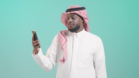 متابعة البرامج المفضلة, عرض شاشة هاتف محمول, دمج التقنية والتكنولوجيا بالحياة اليومية، بورتريه لرجل أسمر عربي خليجي سعودي مبتسم يرتدي الثوب التقليدي و الشماغ و يشير باصبع السبابة الي الجوال, استخدام تطبيقات وخدمات الموبايل, خلفية زرقاء
