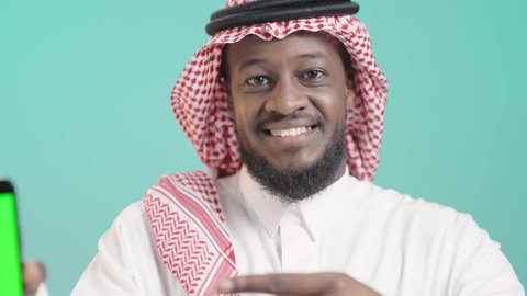 عرض شاشة هاتف محمول, دمج التقنية والتكنولوجيا بالحياة اليومية، بورتريه لرجل أسمر عربي خليجي سعودي مبتسم يرتدي الثوب التقليدي و الشماغ و يشير باصبع السبابة الي الجوال, استخدام تطبيقات وخدمات الموبايل, خلفية زرقاء