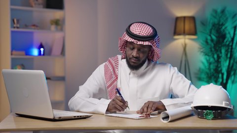 متابعة آخر مستجدات التكنولوجيا الهندسية, التعديل على خطط المشروع ومتابعة العمل, مفهوم الهندسة الإنشائية, بورتريه لرجل أسمر عربي خليجي سعودي يرتدي الثوب التقليدي و الشماغ و بجانبه خوذة الحماية يقوم بالكتابة و استخدام الحاسوب المحمول