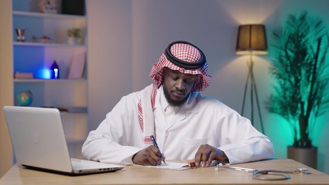 تدوين و تسجيل الملاحظات المعروضة على اللابتوب, العمل في قطاع الصحة والرعاية، بورتريه لرجل أسمر عربي خليجي سعودي يرتدي الثوب التقليدي و الشماغ و المعطف الطبي يقوم بالكتابة و استخدام الحاسوب المحمول, متابعة حالات المرضى عبر الاجهزة و التقنيات الحديثة