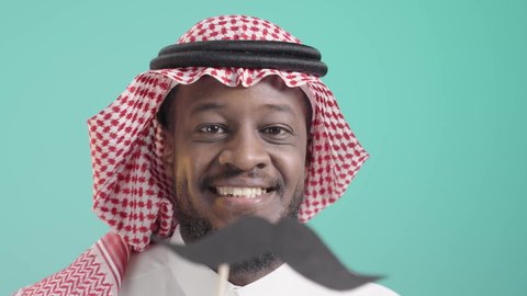 النظر إلى الكاميرا بإيماءات الثقة والاعتزاز بالنفس، مفهوم القوة والرجولة، بورتريه مقرب لشاب أسمر عربي خليجي سعودي يرتدي الثوب التقليدي والشماغ يمسك بيده قصاصة ورقية على شكل شارب، ذكرى اليوم العالمي للرجل ١٩نوفمبر، الاهتمام بالصحة الجسدية للذكور، خلفية زرقاء