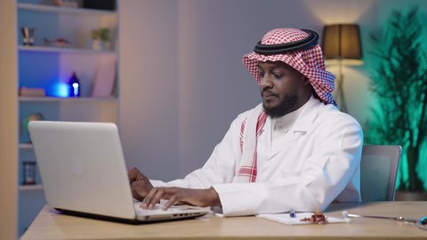 استخدام الأجهزة التقنية الحديثة في الطب، تقديم الاستشارات الطبية عن طريق الإنترنت، العمل في قطاع الصحة والرعاية، بورتريه لرجل أسمر عربي خليجي سعودي يرتدي الثوب التقليدي و الشماغ و المعطف الطبي و يقوم باستخدام اللابتوب