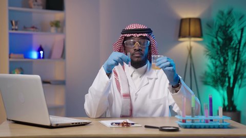 مفهوم الكيمياء الحيوية, استعمال الأنابيب الزجاجية المعقمة لتحليل العينات المختلفة، مختبر التحاليل والفحوصات الطبية، بورتريه لرجل أسمر عربي خليجي سعودي يرتدي الثوب التقليدي و الشماغ و المعطف الطبي ونظارات الحماية و القفازات الطبية ينظر لأنبوب الاختبار بإيماءات التركيز والدقة و بجانبه اللابتوب