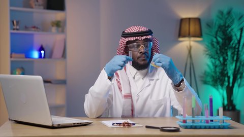 استعمال الأنابيب الزجاجية المعقمة لتحليل العينات المختلفة، مختبر التحاليل والفحوصات الطبية، بورتريه لرجل أسمر عربي خليجي سعودي يرتدي الثوب التقليدي و الشماغ و المعطف الطبي ونظارات الحماية و القفازات الطبية ينظر لأنبوب الاختبار بإيماءات التركيز والدقة و بجانبه اللابتوب