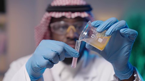 إجراء التجارب الكيميائية باستخدام أنابيب الزجاجية المعقمة, البحث والتطور والقيام باختبارات علمية, بورتريه مقرب ليد رجل أسمر عربي خليجي سعودي يرتدي الثوب التقليدي و الشماغ و المعطف الطبي ونظارات الحماية و القفازات الطبية و يمسك أنبوب الاختبار