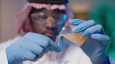 مختبر التحاليل والفحوصات الطبية، إجراء التجارب الكيميائية باستخدام أنابيب الزجاجية المعقمة, البحث والتطور والقيام باختبارات علمية, بورتريه مقرب ليد رجل أسمر عربي خليجي سعودي يرتدي الثوب التقليدي و الشماغ و المعطف الطبي ونظارات الحماية و القفازات الطبية و يمسك أنبوب الاختبار