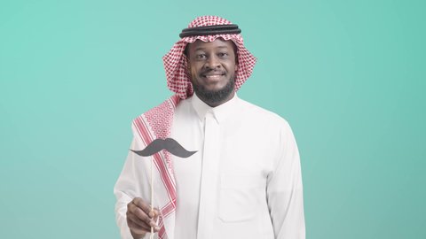 الإحتفال بالمناسبات العالمية، اليوم العالمي للرجل 19 نوفمبر، بورتريه لرجل أسمر عربي خليجي سعودي يرتدي الثوب التقليدي والشماغ يمسك بيده عود خشبي مع قصاصة ورقية على شكل شارب، الإهتمام بصحة الرجال، ايماءات السعادة والسرور, خلفية زرقاء