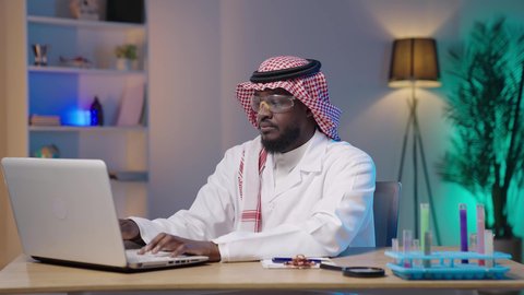 دمج التقنية الحديثة والمتطورة بالمجال الطبي, إجراء التجارب والبحوث العلمية, مفهوم التكنولوجيا الحيوية, مختبر التحاليل والفحوصات الطبية، بورتريه لرجل أسمر عربي خليجي سعودي يرتدي الثوب التقليدي و الشماغ و المعطف الطبي ونظارات الحماية يقوم بالنقر على جهاز اللابتوب
