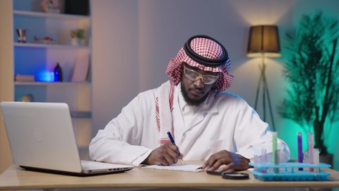 تدوين النتائج والملاحظات, إجراء التجارب والبحوث العلمية, مفهوم التكنولوجيا الحيوية, مختبر التحاليل والفحوصات الطبية، بورتريه لرجل أسمر عربي خليجي سعودي يرتدي الثوب التقليدي و الشماغ و المعطف الطبي ونظارات الحماية يقوم بالكتابة على ورقة و بجانبه جهاز اللابتوب