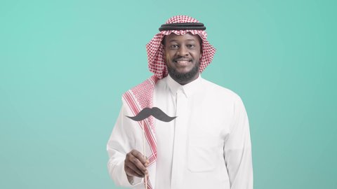 اليوم العالمي للرجل 19 نوفمبر، الإحتفال بالمناسبات العالمية، بورتريه لرجل أسمر عربي خليجي سعودي يرتدي الثوب التقليدي والشماغ  يمسك بيده عود خشبي مع قصاصة ورقية على شكل شارب، الإهتمام بصحة الرجال، ايماءات السعادة والسرور, خلفية زرقاء