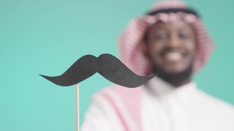 ذكرى اليوم العالمي للرجل 19نوفمبر، بورتريه مقرب للرجل عربي خليجي سعودي يرتدي الثوب التقليدي والشماغ يمسك بيده عود خشبي مع قصاصة ورقية على شكل شارب و يقربها للكاميرا, الاهتمام بالصحة الجسدية للذكور، خلفية زرقاء