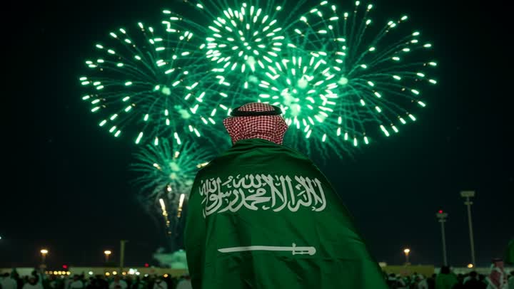 Watching fireworks in the evening, a back view of a Saudi Arabian Gulf man wearing a shemagh and placing the flag of the Kingdom of Saudi Arabia on his shoulders, celebrating national occasions and holidays, Flag Day March 11, Saudi National Day September 23, commemorating the 95th anniversary of Saudi Arabia