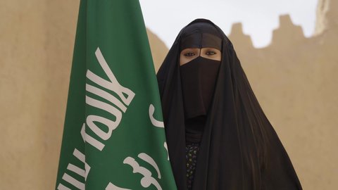 الاحتفال باليوم الوطني 23 سبتمبر, ذكرى يوم التأسيس 22 فبراير, سيدة عربية خليجية سعودية ترتدي العباءة السوداء والبرقع وتقف بجانب العلم, يوم العلم السعودي 11 مارس, التفاخر بالإنتماء للوطن, ارتداء الأزياء الشعبية التقليدية في المناسبة الوطنية