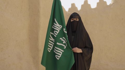 Celebrating National Day on September 23, Founding Day on February 22, a Saudi Arabian Gulf woman wearing a black abaya and burqa standing next to the flag, Saudi Flag Day on March 11, pride in belonging to the homeland, wearing traditional folk costumes on the national occasion