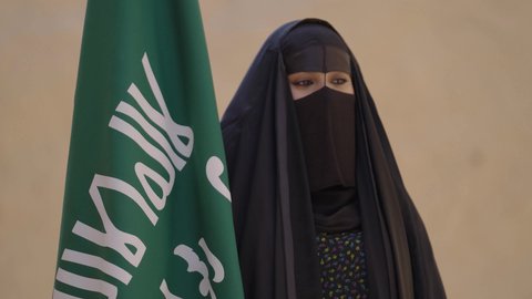 Pride in belonging to the homeland, wearing traditional folk costumes on a national occasion, celebrating National Day on September 23, Founding Day on February 22, a Saudi Arabian Gulf woman wearing a black abaya and burqa standing next to the flag, Saudi Flag Day on March 11