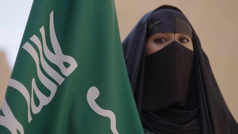Pride in belonging to the homeland, wearing traditional folk costumes on a national occasion, celebrating National Day on September 23, Founding Day on February 22, a Saudi Arabian Gulf woman wearing a black abaya and burqa standing next to the flag, Saudi Flag Day on March 11