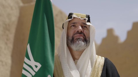 ارتداء الزي الشعبي في مناسبة وطنية, الاحتفال بيوم العلم السعودي11مارس، يوم بدينا22 فبراير, مفهوم حب الوطن والانتماء اليه, ذكرى تأسيس الدولة السعودية الأولى1727م, رجل عربي خليجي سعودي يرتدي البشت والغترة يقف في منزل طيني ويمسك علم يوم التأسيس بتعابير الشموخ