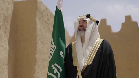 Wearing traditional costume on a national occasion, celebrating Saudi Flag Day March 11, our beginning day February 22, the concept of patriotism and belonging to the homeland, the anniversary of the founding of the first Saudi state 1727 AD, a Saudi Gulf Arab man wearing a bisht and a ghutra standing in a mud house and holding the Founding Day flag with expressions of pride