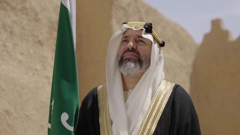 ارتداء الزي الشعبي في مناسبة وطنية, الاحتفال بيوم العلم السعودي11مارس، يوم بدينا22 فبراير, مفهوم حب الوطن والانتماء اليه, ذكرى تأسيس الدولة السعودية الأولى1727م, رجل عربي خليجي سعودي يرتدي البشت والغترة يقف في منزل طيني ويمسك علم يوم التأسيس بتعابير الشموخ