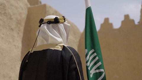 ارتداء الزي الشعبي في مناسبة وطنية, الاحتفال بيوم العلم السعودي11مارس، يوم بدينا22 فبراير, مفهوم حب الوطن والانتماء اليه, ذكرى تأسيس الدولة السعودية الأولى1727م, رجل عربي خليجي سعودي يرتدي البشت والغترة يقف في منزل طيني ويمسك علم يوم التأسيس بتعابير الشموخ