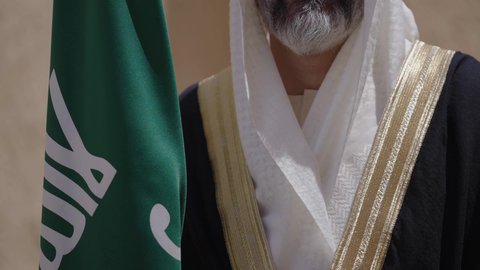 ارتداء الزي الشعبي في مناسبة وطنية, الاحتفال بيوم العلم السعودي11مارس، يوم بدينا22 فبراير, مفهوم حب الوطن والانتماء اليه, ذكرى تأسيس الدولة السعودية الأولى1727م, رجل عربي خليجي سعودي يرتدي البشت والغترة يقف في منزل طيني ويمسك علم يوم التأسيس بتعابير الشموخ