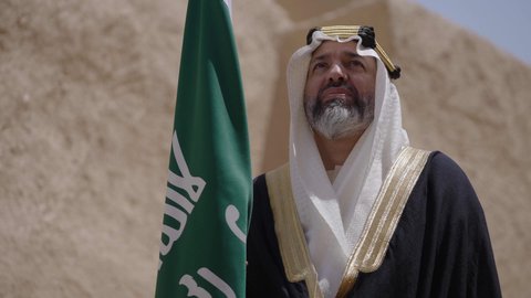 الاحتفال بيوم العلم السعودي11مارس، يوم بدينا22 فبراير, مفهوم حب الوطن والانتماء اليه, ذكرى تأسيس الدولة السعودية الأولى1727م, رجل عربي خليجي سعودي يرتدي البشت والغترة يقف في منزل طيني ويمسك علم يوم التأسيس بتعابير الشموخ، ارتداء الزي الشعبي في مناسبة وطنية
