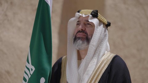 الاحتفال بيوم العلم السعودي11مارس، يوم بدينا22 فبراير, مفهوم حب الوطن والانتماء اليه, ذكرى تأسيس الدولة السعودية الأولى1727م, رجل عربي خليجي سعودي يرتدي البشت والغترة يقف في منزل طيني ويمسك علم يوم التأسيس بتعابير الشموخ، ارتداء الزي الشعبي في مناسبة وطنية