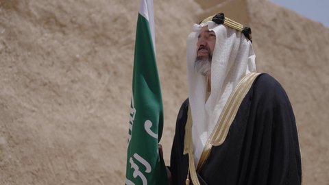 Celebrating Saudi Flag Day, March 11, our beginning day, February 22, the concept of patriotism and belonging to the homeland, the anniversary of the founding of the first Saudi state 1727 AD, a Saudi Gulf Arab man wearing a bisht and ghutra standing in a mud house and holding the founding day flag with expressions of pride, wearing traditional dress on a national occasion