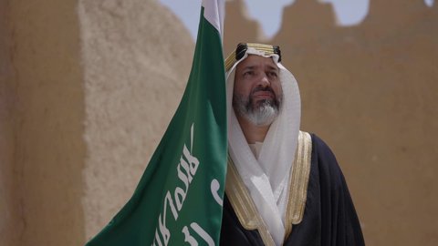 Celebrating Saudi Flag Day, March 11, our beginning day, February 22, the concept of patriotism and belonging to the homeland, the anniversary of the founding of the first Saudi state 1727 AD, a Saudi Gulf Arab man wearing a bisht and ghutra standing in a mud house and holding the founding day flag with expressions of pride, wearing traditional dress on a national occasion