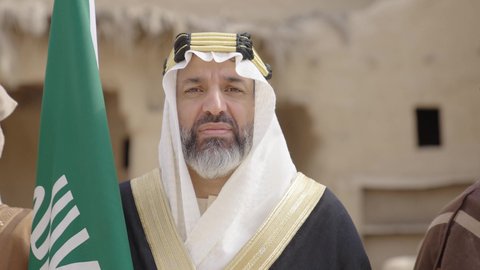 الاحتفال بيوم العلم السعودي11مارس، يوم بدينا22 فبراير, مفهوم حب الوطن والانتماء اليه, ذكرى تأسيس الدولة السعودية الأولى1727م, رجل عربي خليجي سعودي يرتدي البشت والغترة يقف في منزل طيني ويمسك علم يوم التأسيس بتعابير الشموخ، ارتداء الزي الشعبي في مناسبة وطنية