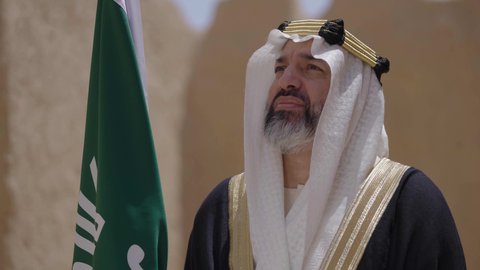 The day we began, February 22, the concept of patriotism and belonging to the homeland, the anniversary of the founding of the first Saudi state 1727 AD, a Saudi Gulf Arab man wearing a bisht and ghutra standing in a mud house and holding the Founding Day flag with expressions of pride, wearing traditional dress on a national occasion, celebrating Saudi Flag Day, March 11