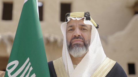 يوم بدينا22 فبراير, مفهوم حب الوطن والانتماء اليه, ذكرى تأسيس الدولة السعودية الأولى1727م, رجل عربي خليجي سعودي يرتدي البشت والغترة يقف في منزل طيني ويمسك علم يوم التأسيس بتعابير الشموخ، ارتداء الزي الشعبي في مناسبة وطنية, الاحتفال بيوم العلم السعودي11مارس