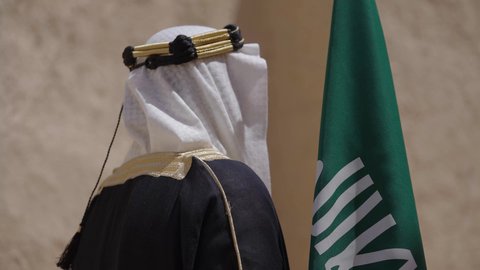 يوم بدينا22 فبراير, مفهوم حب الوطن والانتماء اليه, ذكرى تأسيس الدولة السعودية الأولى1727م, رجل عربي خليجي سعودي يرتدي البشت والغترة يقف في منزل طيني ويمسك علم يوم التأسيس بتعابير الشموخ، ارتداء الزي الشعبي في مناسبة وطنية, الاحتفال بيوم العلم السعودي11مارس