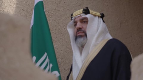 يوم بدينا22 فبراير, مفهوم حب الوطن والانتماء اليه, ذكرى تأسيس الدولة السعودية الأولى1727م, رجل عربي خليجي سعودي يرتدي البشت والغترة يقف في منزل طيني ويمسك علم يوم التأسيس بتعابير الشموخ، ارتداء الزي الشعبي في مناسبة وطنية, الاحتفال بيوم العلم السعودي11مارس