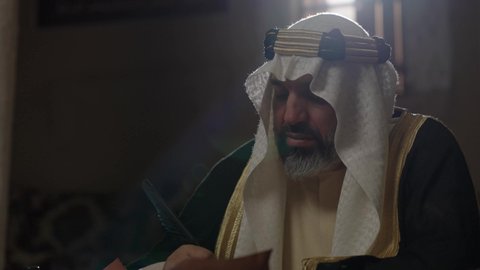 Wearing traditional costume on the national occasion, a close-up of a Saudi Arabian Gulf man wearing a ghutra, golden agal, and bisht holding a pen in his hand, writing on a historical leather manuscript, heritage letters and correspondence for the establishment of the first state in 1727 AD, the day we began, February 22, pride in the authenticity of the Kingdom