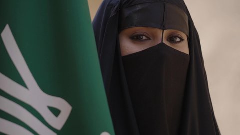 التفاخر بالإنتماء للوطن, ارتداء الأزياء الشعبية التقليدية في المناسبة الوطنية، الاحتفال باليوم الوطني 23 سبتمبر, ذكرى يوم التأسيس 22 فبراير, سيدة عربية خليجية سعودية ترتدي العباءة السوداء والبرقع وتقف بجانب العلم, يوم العلم السعودي 11 مارس