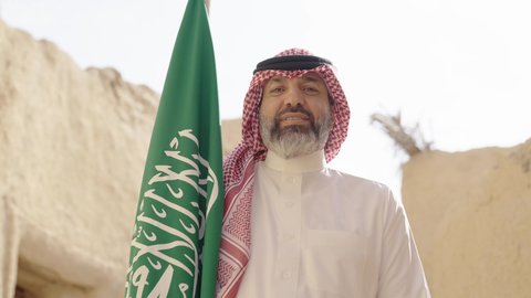 The concept of patriotism and belonging to the homeland, the anniversary of the founding of the first Saudi state 1727 AD, an elderly Saudi Gulf Arab man wearing a traditional shemagh and thobe holding the national flag in his hand to celebrate Flag Day, March 11, a walk in an old traditional popular place, the day of our beginning, February 22