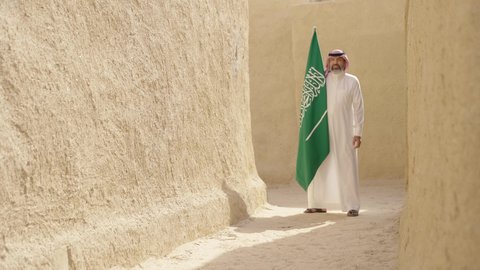 The concept of patriotism and belonging to the homeland, the anniversary of the founding of the first Saudi state 1727 AD, an elderly Saudi Gulf Arab man wearing a traditional shemagh and thobe holding the national flag in his hand to celebrate Flag Day, March 11, a walk in an old traditional popular place, the day of our beginning, February 22