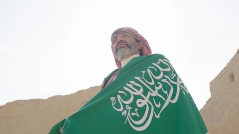 The concept of patriotism and belonging to the homeland, the anniversary of the founding of the first Saudi state 1727 AD, an elderly Saudi Gulf Arab man wearing a traditional shemagh and thobe holding the national flag in his hand to celebrate Flag Day, March 11, a walk in an old traditional popular place, the day of our beginning, February 22
