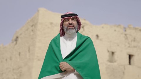 مفهوم حب الوطن والانتماء اليه, ذكرى تأسيس الدولة السعودية الأولى1727 م, رجل كبير بالسن عربي خليجي سعودي يرتدي الشماغ والثوب التقليدي يمسك بيده راية الوطن للاحتفال بيوم العلم 11 مارس، التجول في مكان شعبي تقليدي قديم، يوم بدينا 22 فبراير