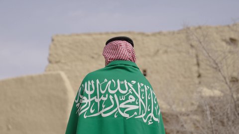 حب الوطن والانتماء اليه, ذكرى تأسيس الدولة السعودية الأولى1727 م, صورة من الخلف لرجل كبير بالسن عربي خليجي سعودي يرتدي الشماغ والثوب التقليدي يمسك بيده راية الوطن للاحتفال بيوم العلم 11 مارس، التجول في مكان شعبي تقليدي قديم، يوم بدينا 22 فبراير