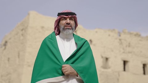 The concept of patriotism and belonging to the homeland, the anniversary of the founding of the first Saudi state 1727 AD, an elderly Saudi Gulf Arab man wearing a traditional shemagh and thobe holding the national flag in his hand to celebrate Flag Day, March 11, a walk in an old traditional popular place, the day of our beginning, February 22